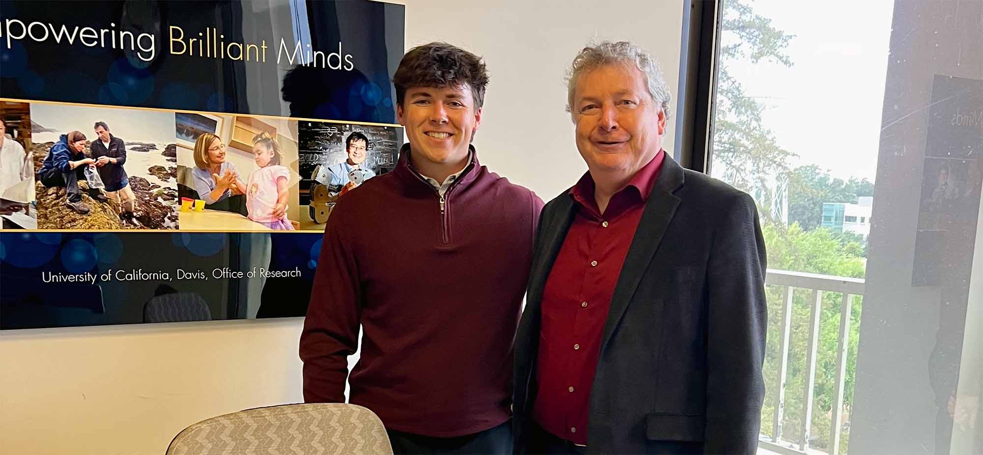 Two men smiling by an office window; younger in maroon sweater, older in blazer