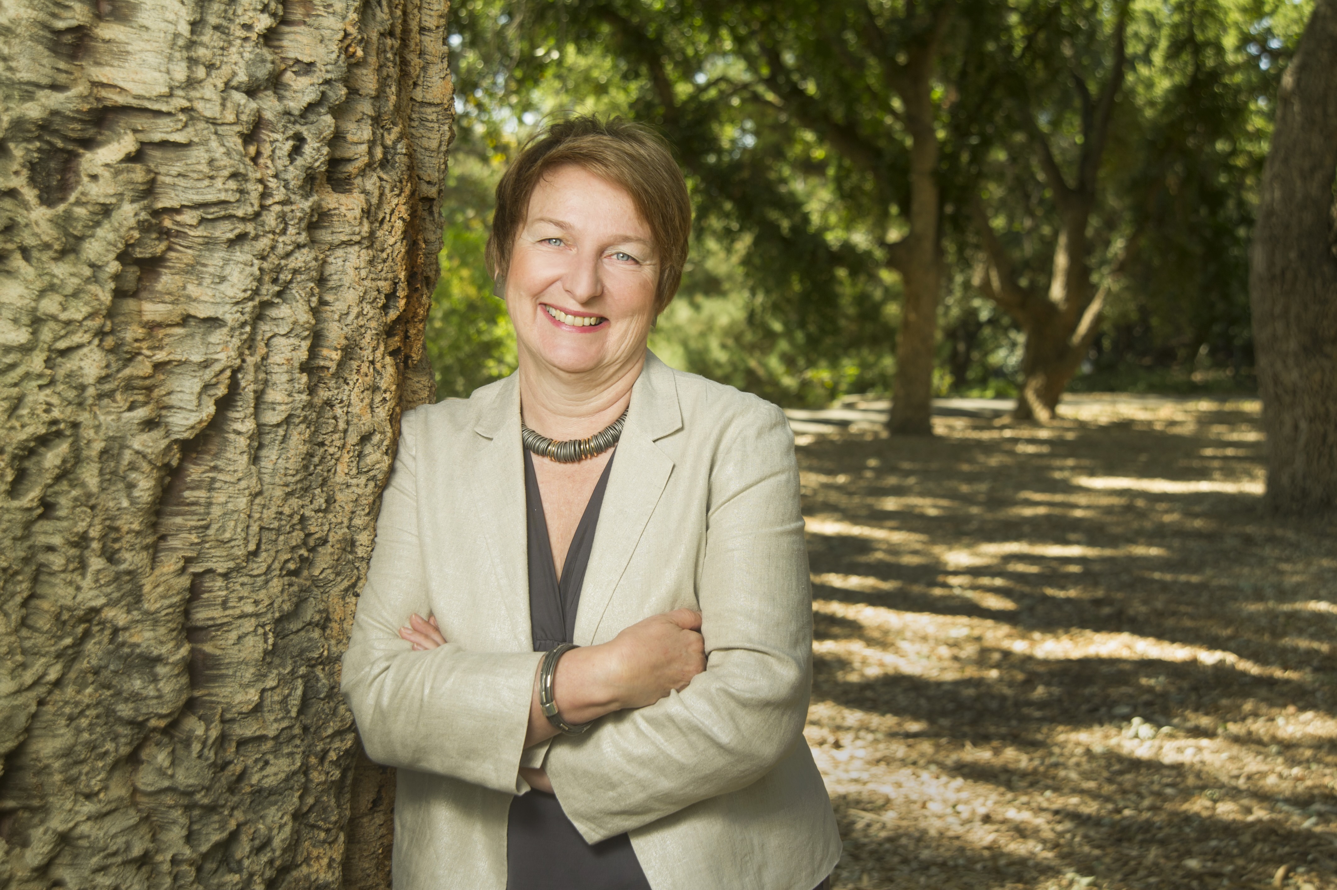 Smiling woman in light blazer leaning against a tree in a sunlit park 
