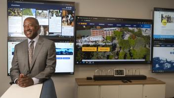 Chancellor Gary May stands in front of a group of large display screens.