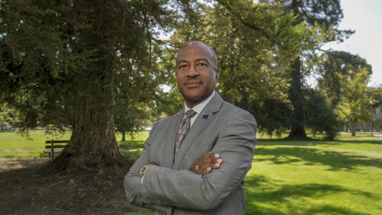 Chancellor May wears a grey suit with his arms crossed on his body, standing outside in front of a green lawn and oak tree that appears in the background.