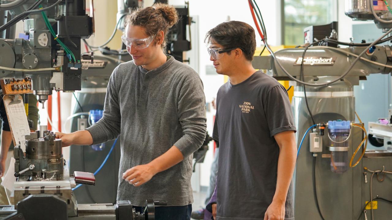 A male student wearing a grey long-sleeve shirt and protective clear goggles operates machinery in a student workshop, while a male student stands to the right to watch wearing a blue t-shirt and clear protective goggles.