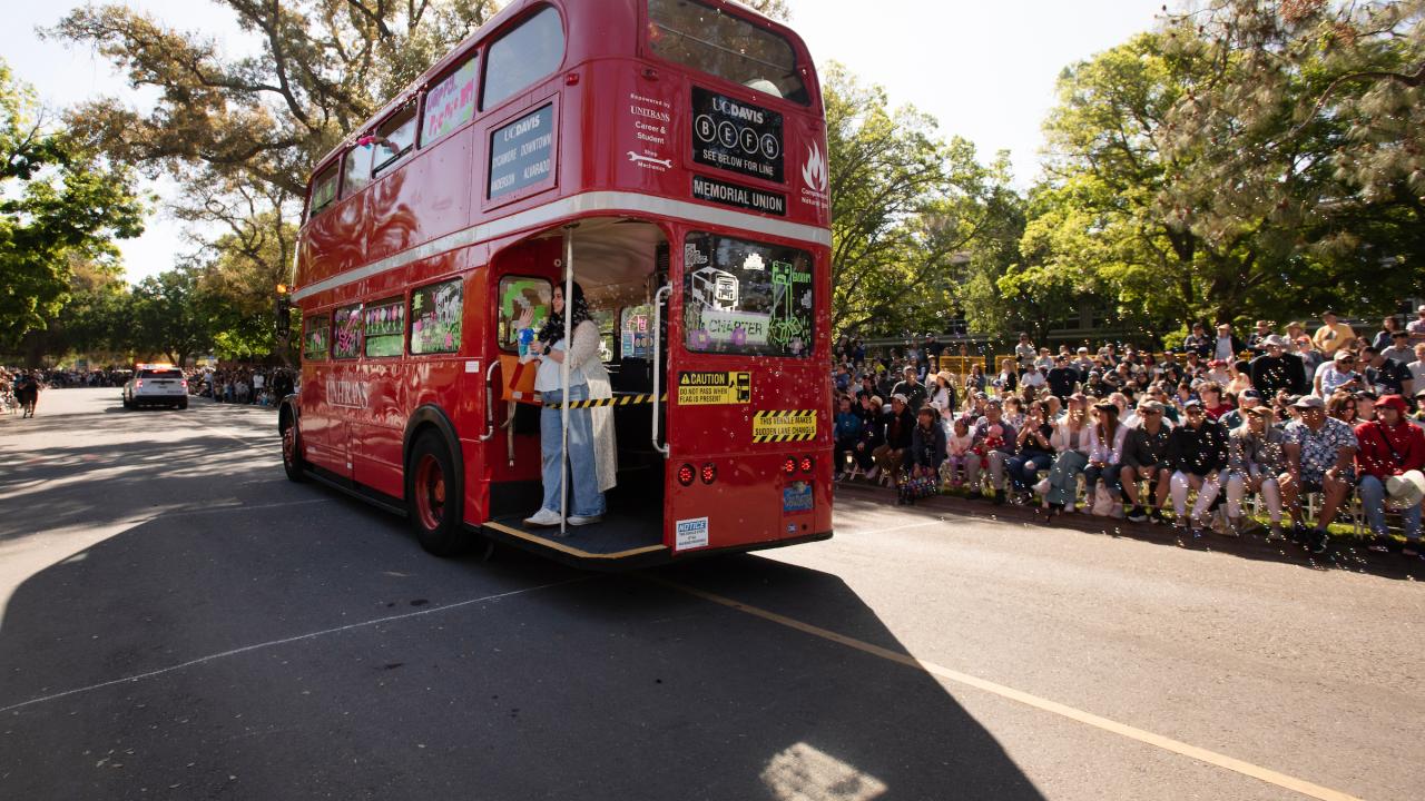 A red double-decker bus drives down the street in front of a parade crowd.