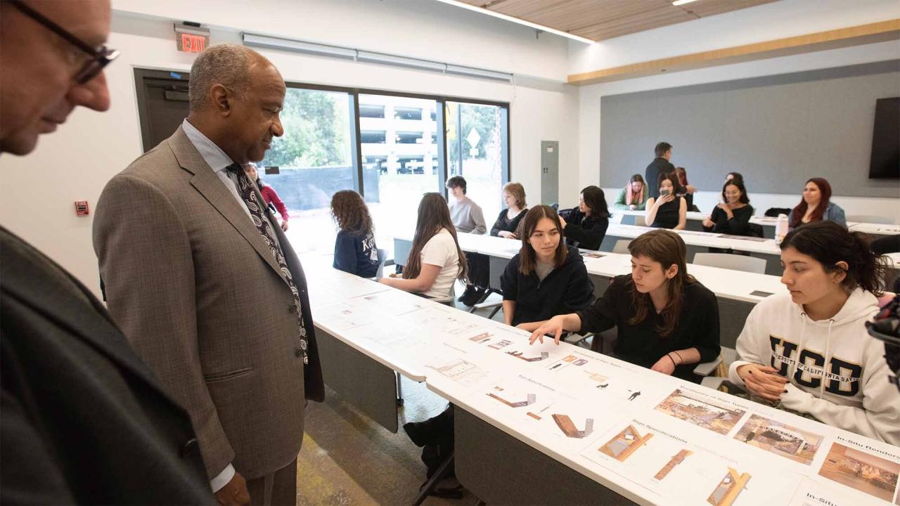 A group of students dressed in black hoodies and a white sweater show Chancellor May dressed in a brown blazer jacket informational graphics on a white table. 