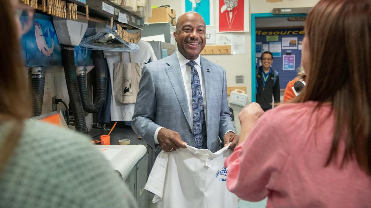 "Chancellor May, in suit and tie, smiles while holding screen-printed T-shirt and being interviewed live for morning TV show"