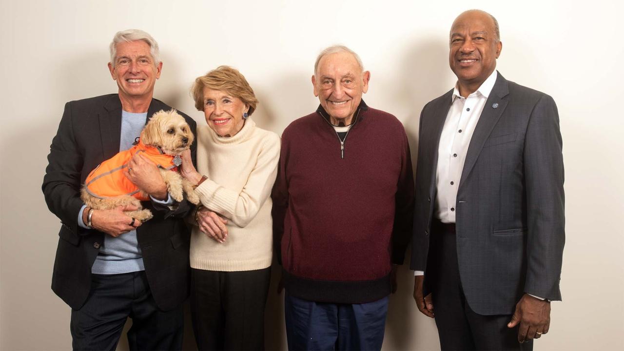Four smiling individuals stand together, with one holding a small dog in a bright orange vest.