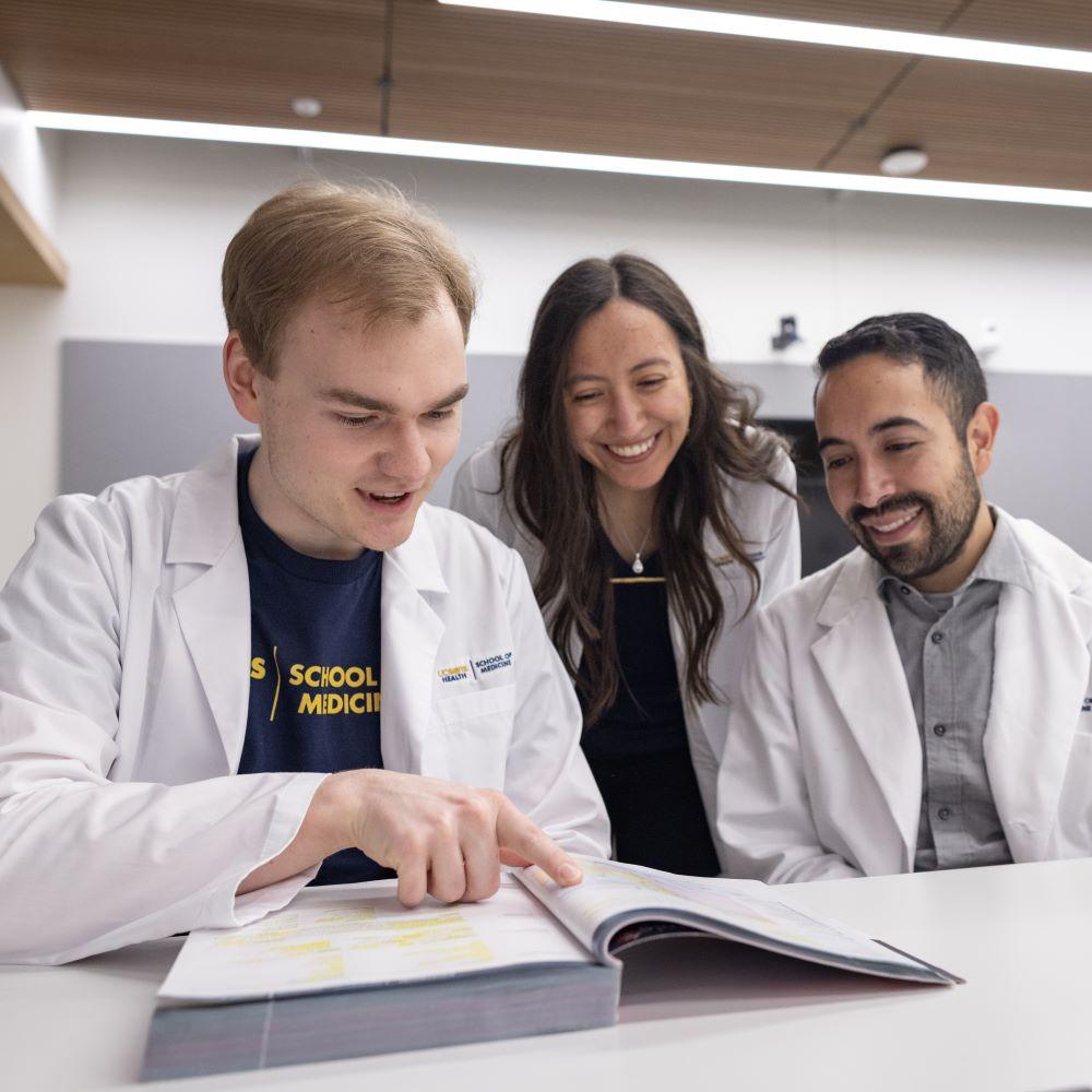 Three medical trainees in white lab coats review a textbook together at a table, discussing content in a classroom or study space.on training session in a classroom setting.