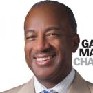 Chancellor Gary May smiling, looking at camera on white background. Words "Gary May Chancell-ing" is overlaid on the white background.