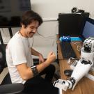 A person sits at a desk with a robotic arm, smiling and making a fist.