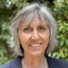 Smiling older woman with gray hair in navy blazer, headshot photo, blurred greenery background