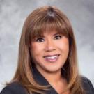 Professional headshot photograph of smiling woman with shoulder-length brown hair, gray background