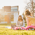 Two students wearing face masks sitting on picnic blanket outside of residence hall